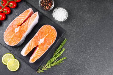 Siyah taş yüzeyinde iki taze çiğ somon bifteği ve besin maddeleri. Domates, limon, biberiye, tuz ve biber. Sağlıklı beslenme, deniz ürünleri, yemek pişirme, tarif ve menü kavramları. Üst görünüm, alanı kopyala