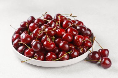 Taze olgun kırmızı kiraz yığını. Arka planda beyaz tabakta sapları var. Doğal yaz meyvesi, sağlıklı organik gıda, hasat ve mevsimsel kavramlar..