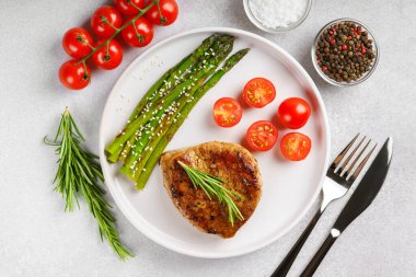 Lezzetli ızgara biftek, yeşil kuşkonmaz ve beyaz tabakta vişneli domates, çatal bıçak ve baharatla servis ediliyor. Yemek menüsü, diyet ya da mutfak tasarımı için mükemmel..