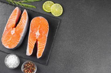 Siyah taş yüzeyinde iki taze çiğ somon bifteği ve besin maddeleri. Domates, limon, biberiye, tuz ve biber. Sağlıklı beslenme, deniz ürünleri, yemek pişirme, tarif ve menü kavramları. Üst görünüm, alanı kopyala