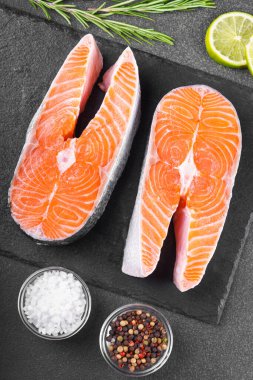 İki taze çiğ somon ya da besin maddeleri içeren siyah taş yüzeyde alabalık bifteği. Domates, limon, biberiye, tuz ve biber. Sağlıklı beslenme, deniz ürünleri, yemek pişirme, tarif ve menü kavramları.