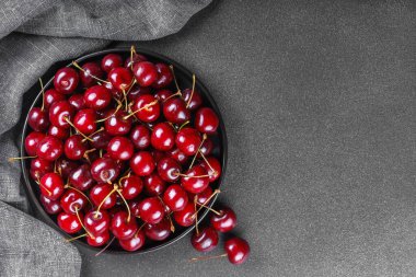 Koyu dokulu arka planda gri keten peçeteye yerleştirilmiş olgun kırmızı kirazla dolu siyah tabağı kapat. Lezzetli yemekler, sulu yaz meyveleri, sağlıklı yiyecekler. Üst görünüm, alanı kopyala
