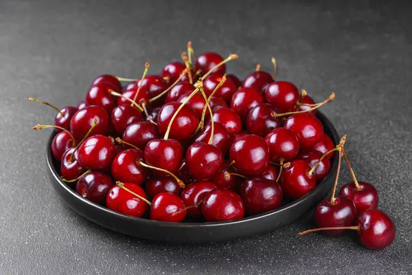 Koyu gri desenli arka plana yerleştirilmiş olgun kırmızı kirazla dolu siyah tabağı kapat. Lezzetli yemekler, sulu yaz meyveleri ve sağlıklı yiyecekler..