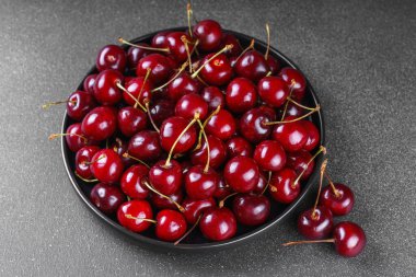 Koyu gri desenli arka plana yerleştirilmiş olgun kırmızı kirazla dolu siyah tabağı kapat. Lezzetli yemekler, sulu yaz meyveleri ve sağlıklı yiyecekler..
