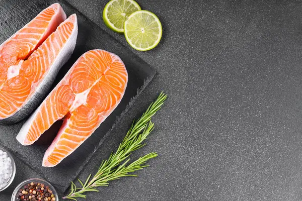 İki taze çiğ somon ya da besin maddeleri içeren siyah taş yüzeyde alabalık bifteği. Domates, limon, biberiye, tuz ve biber. Sağlıklı beslenme, deniz ürünleri, yemek pişirme, yemek tarifi ve menü. Üst görünüm, alanı kopyala