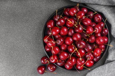Koyu dokulu arka planda gri keten peçeteye yerleştirilmiş olgun kırmızı kirazla dolu siyah tabağı kapat. Lezzetli yemekler, sulu yaz meyveleri, sağlıklı yiyecekler. Üst görünüm, alanı kopyala