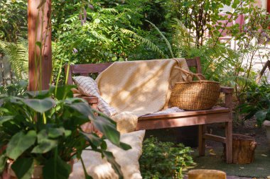 Yumuşak sarı battaniyeli, çizgili yastıklı ahşap bir bank ve saman sepeti. Sıcak yaz güneşi altında yemyeşil bahçede dinlenmek için yer var..