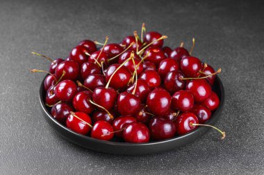 Koyu gri desenli arka plana yerleştirilmiş olgun kırmızı kirazla dolu siyah tabağı kapat. Lezzetli yemekler, sulu yaz meyveleri ve sağlıklı yiyecekler..
