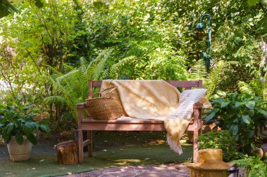 Yumuşak sarı battaniyeli, çizgili yastıklı ahşap bir bank ve saman sepeti. Sıcak yaz güneşi altında yemyeşil bahçede dinlenmek için yer var..