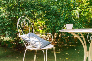 Eski, dövülmüş beyaz metal sandalyeli ve rahat yastıklı huzurlu bir bahçe kafesi. Beyaz kahve fincanı olan yuvarlak masanın yanında yemyeşil arka planda, yaz gününde rahatlatıcı atmosfer..