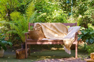 Yumuşak sarı battaniyeli, çizgili yastıklı ahşap bir bank ve saman sepeti. Sıcak yaz güneşi altında yemyeşil bahçede dinlenmek için yer var..