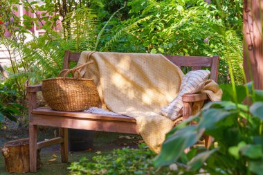 Yumuşak sarı battaniyeli, çizgili yastıklı ahşap bir bank ve saman sepeti. Sıcak yaz güneşi altında yemyeşil bahçede dinlenmek için yer var..