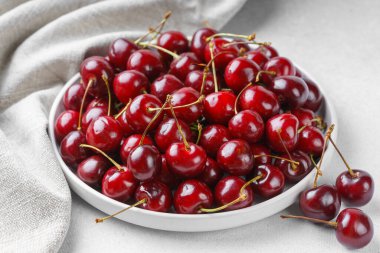 Açık arkaplanda gri keten peçeteye yerleştirilmiş olgun kırmızı kirazla dolu beyaz tabağı kapat. Lezzetli yemekler, sulu yaz meyveleri ve sağlıklı yiyecekler..