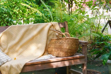 Yumuşak sarı battaniyeli, çizgili yastıklı ahşap bir bank ve saman sepeti. Sıcak yaz güneşi altında yemyeşil bahçede dinlenmek için yer var..