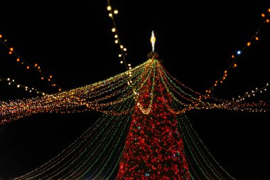 Parlak ışıklandırılmış büyük Noel ağacı. Parlak ışıklar ve gece süsleriyle süslenmiş çelenklerle süslenmiş. Bayram atmosferi yaratıyor. Noel ve Yeni Yıl Tatil Fuarı.