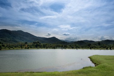 Taman Tasik Milenyum Gölü, Kuala Kubu Bharu, Malezya 'nın manzarası - Taman Tasik Milenyumu' ndaki dingin göl, yemyeşil bir kıyı şeridi ve parçalı bulutlu bir gökyüzünün altında ormanlık tepelerin yer aldığı sakin bir manzara. 