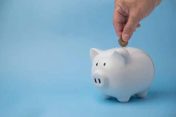 Money management and financial success. Man hand putting coins in a white piggy bank for wealth accumulation and financial independence and investment on background.