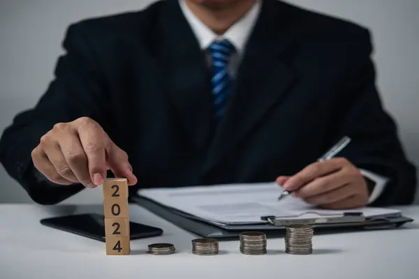 Businessman hand stacking wooden cube 2024 and coins stacking on desk. business Money Saving budget planning investment and Financial and tax concept.