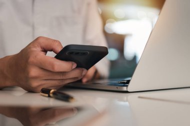 Businessman hand holding mobile smartphone on a table with a laptop at office..	