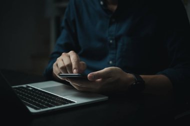 Bir adam dizüstü bilgisayarla masada otururken cep telefonunu kullanıyor. Adam telefonuna odaklanmış, muhtemelen internete mesaj atıyor ya da internette geziniyor. Üretkenlik ve çoklu görev kavramı