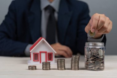 İş adamı, küçük ev modeliyle parayı kavanoza koyuyor. Gayrimenkul yatırımı için para biriktirme kavramı. Mali planlama, ev sahipliği, ipotek ve bankacılık.