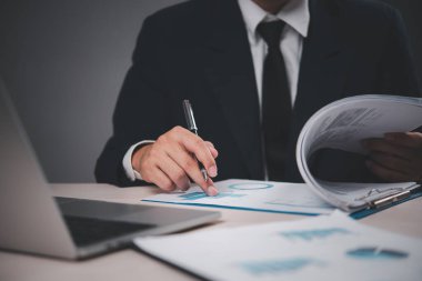 Takım elbiseli bir işadamı masa başında belgeleri analiz ediyor. El ele tutuşup çizelgeleri gösteriyorlar. Profesyonel, finansal analize odaklı..