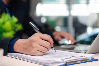 İş adamı ofis ortamında dizüstü bilgisayarla çalışan belgeleri imzalıyor. Bilgisayarın çalışma alanının yanında kağıt belgeler üzerine kalem ile yönetici el yazısının yakın görüntüsü.
