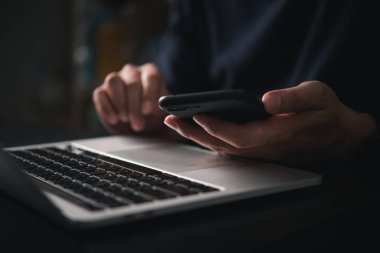 Ofiste ya da ev ortamında dizüstü bilgisayarda yazarken elinde akıllı telefon tutan adam, çevrimiçi işleri, iş görevlerini, e-postaları ve teknolojiyi kullanarak internet iletişimini temsil ediyor.