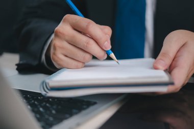 Bir dizüstü bilgisayar üzerinde çalışırken kalemle deftere profesyonel bir yazı yazmak. İmaj, ofisteki verimliliği ve iş planlamasını simgeliyor..