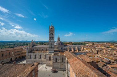 Siena Katedrali 'nin ikonik çizgili çan kulesiyle panoramik bir görüntüsü, açık mavi gökyüzünün altındaki tarihi binalarla çevrili..