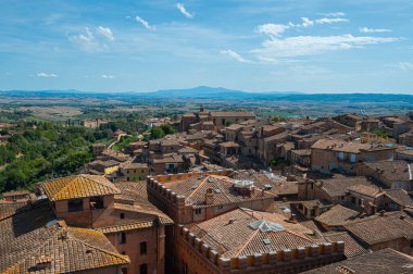 Terasotta çatıları, ortaçağ mimarisi ve açık mavi gökyüzünün altında yuvarlanan tepelerin yer aldığı tarihi İtalyan şehri Siena 'nın panoramik bir görüntüsü..