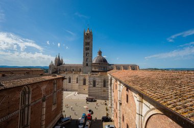 Siena Katedrali 'nin ikonik çizgili çan kulesiyle panoramik bir görüntüsü, açık mavi gökyüzünün altındaki tarihi binalarla çevrili..