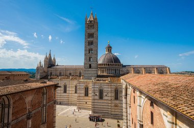 Siena Katedrali 'nin ikonik çizgili çan kulesiyle panoramik bir görüntüsü, açık mavi gökyüzünün altındaki tarihi binalarla çevrili..