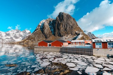 Lofoten 'daki Hamnoy köyünde mavi gökyüzünün altında donmuş bir gölün yanındaki kırmızı kulübelerin manzarası..