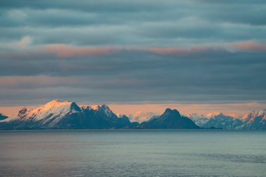 Bulutlu bir gökyüzü altında, kar kaplı dağların sakin manzarası, önplanda sakin bir deniz, günbatımının yumuşak renklerini yansıtıyor, Lofoten, Norveç