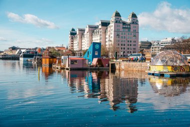 Oslo, Norveç - 13 Nisan 2024: Yüzen renkli evler ve modern binalar açık mavi gökyüzü altında sakin bir limana yansıyor.