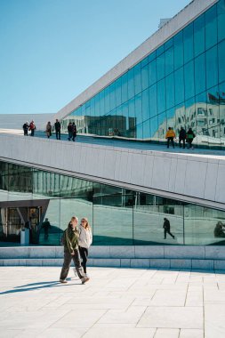 Oslo, Norveç - 13 Nisan 2024: İnsanlar Oslo Opera Binası 'nda yürüyor ve dinleniyor, liman kenarında eğimli çatısı olan modern bir mimari yapı..