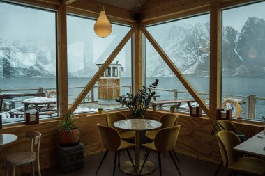 Lofoten, Norveç - 5 Nisan 2024: Karlı bir dağ manzarası ve sakin bir göl içeren geniş pencereli modern mobilyalarla dolu samimi bir kafe.