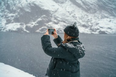 Kışın giyinmiş bir insan, göl kenarındaki karlı bir arazinin fotoğrafını çeker. Nusfjord, Lofoten, Norveç.