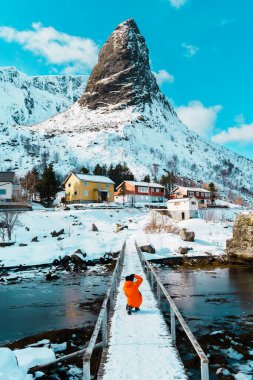 Lofoten, Norveç - 5 Nisan 2024: Turuncu ceketli bir kişi, karlı bir köprüde renkli evlere ve açık mavi gökyüzünün altında yükselen bir dağa doğru yürüyor. Reine, Lofoten.