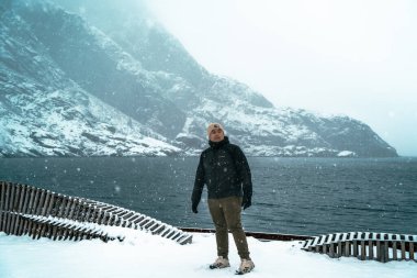 Lofoten, Norveç - APR 6.2024: Arkasında dağlar olan bir göl kenarındaki karlı arazide duran kişi. Nusfjord, Lofoten, Norveç.
