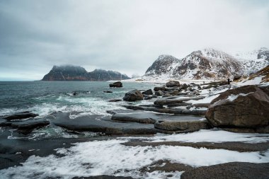 Norveç 'in Lofoten kentindeki Uttakleiv Sahili' nde engebeli kayalar, okyanus dalgaları ve karlı dağ arka planının yer aldığı dramatik kutup sahneleri.