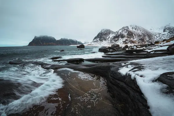 Norveç 'in Lofoten kentindeki Uttakleiv Sahili' nde engebeli kayalar, okyanus dalgaları ve karlı dağ arka planının yer aldığı dramatik kutup sahneleri.