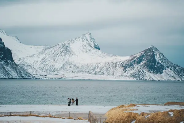 Lofoten, Norveç - APR 7.2024: Norveç 'in Lofoten kentindeki Haukland plajında karla kaplı zirveler, sakin deniz ve keşif yapan ziyaretçilerin bulunduğu manzaralı kış manzarası.