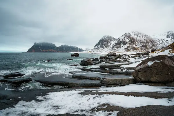 Norveç 'in Lofoten kentindeki Uttakleiv Sahili' nde engebeli kayalar, okyanus dalgaları ve karlı dağ arka planının yer aldığı dramatik kutup sahneleri.