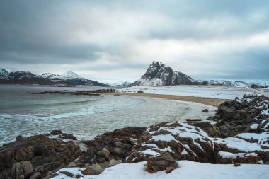 Norveç, Lofoten 'deki Storsandnes Sahili' nde kış manzarası karlı kıyı şeridini, sakin dalgaları ve yüksek engebeli zirveyi gösteriyor..