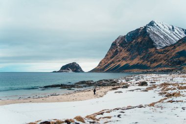 Karlı kıyı şeridi, turkuaz deniz ve bulutlu gökyüzü altında engebeli dağların yer aldığı Vik Sahili, Lofoten, Norveç 'teki huzurlu kış manzarası.