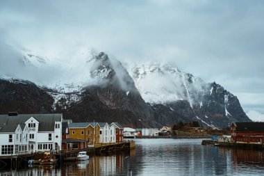 Henningsvaer, Lofoten, Norveç 'in resimli liman manzarası. Balıkçı tekneleri, geleneksel binalar ve karla kaplı dağlar suya yansıyor..