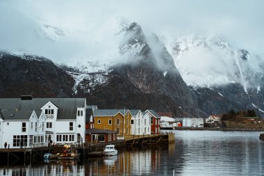 Henningsvaer, Lofoten, Norveç 'in resimli liman manzarası. Balıkçı tekneleri, geleneksel binalar ve karla kaplı dağlar suya yansıyor..
