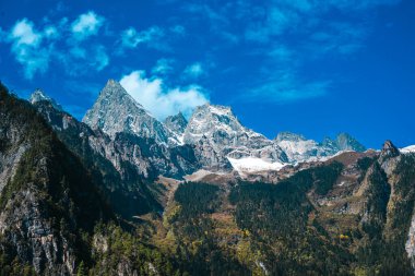 Çin, Balagezong 'da yemyeşil bir ormanı olan engebeli, karla kaplı bir dağ sırasının dramatik manzarası..
